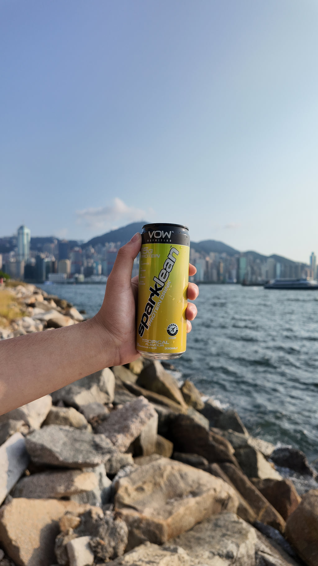 A hand holding Sparklean Tropical by a rocky waterfront with city buildings across the water