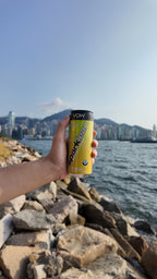 A hand holding Sparklean Tropical by a rocky waterfront with city buildings across the water